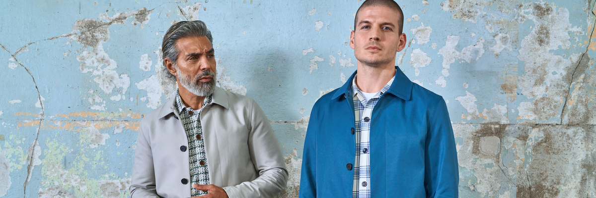 Two men standing in front of a distressed, peeling light blue wall. The man on the left has gray hair and a beard, wearing a light gray Mac jacket over a plaid shirt. The Man on the right wears a blue Mac over a plaid shirt